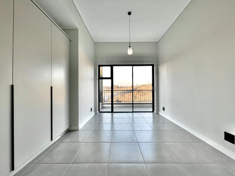 A bright, modern bedroom featuring neutral tiled flooring, ample built-in cupboard space, and a sliding glass door leading to a private balcony.