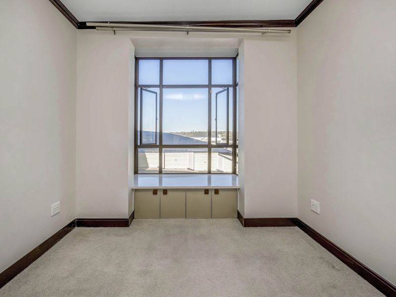 A bright, empty bedroom featuring a large window with built-in storage cabinetry, neutral carpeting, and dark wood trim detailing.
