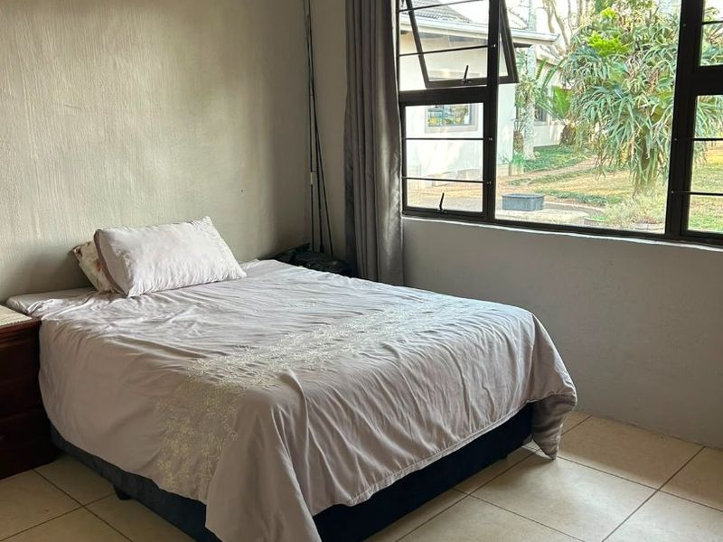 A bedroom featuring tiled flooring, a ceiling fan, and large windows with aluminium frames, offering natural light and a view of the garden.