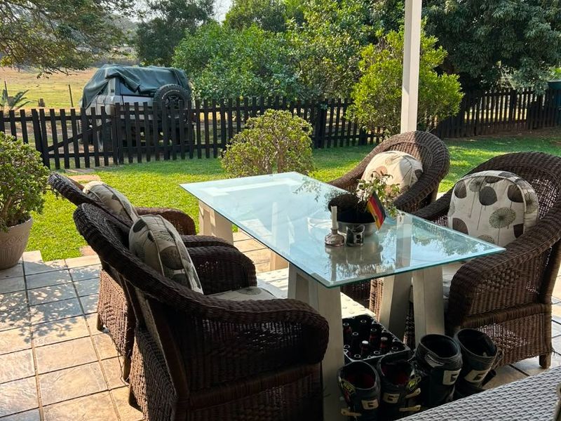 A covered outdoor patio area featuring tiled flooring and a corrugated metal roof, overlooking a private garden and fenced yard.