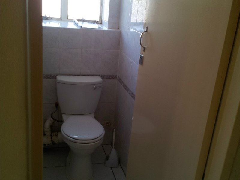 A small, dated bathroom featuring a standard toilet, tiled walls, and tiled flooring, requiring cosmetic updates and plumbing concealment.