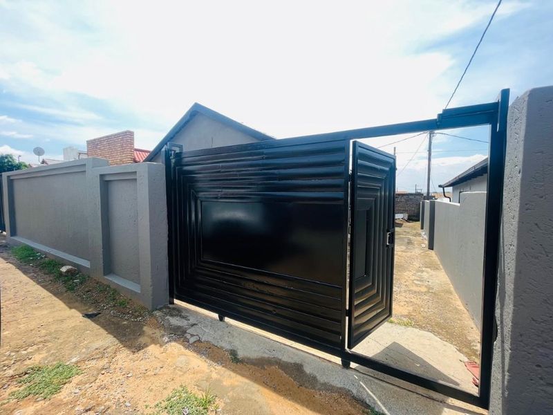 A secure property entrance featuring a modern black metal gate and a solid perimeter wall, leading to an unfinished driveway area.