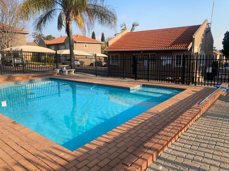 Communal swimming pool area featuring brick paving, metal perimeter fencing, and a nearby clubhouse building with a tiled roof.