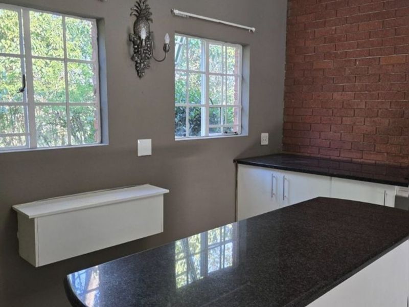 A clean kitchen featuring a central island with granite countertops, an exposed brick feature wall, and neutral grey painted walls.