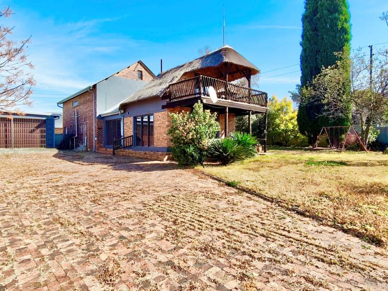 Rear view of a multi-story house featuring a thatched roof, a wooden balcony, and a large brick-paved driveway area.