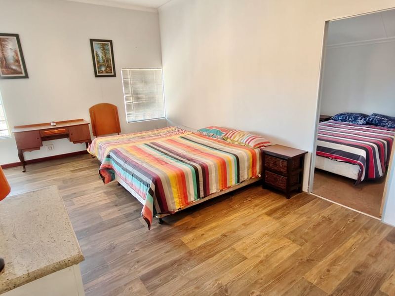 A bedroom featuring laminate flooring and natural light through venetian blinds, with an adjoining room visible through the doorway.