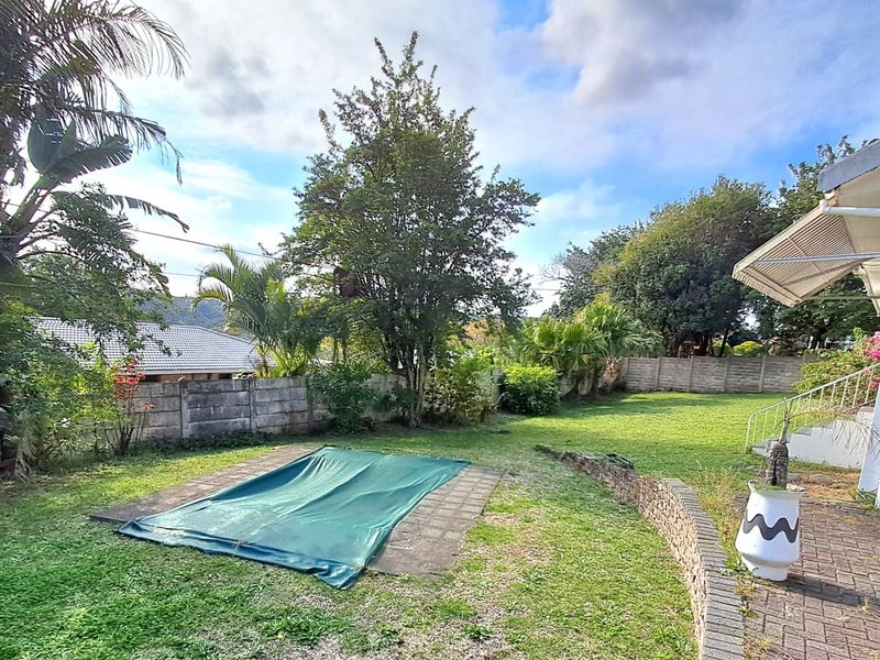 A rear garden area featuring a swimming pool with a cover, paved surround, and a low stone retaining wall bordering the lawn.