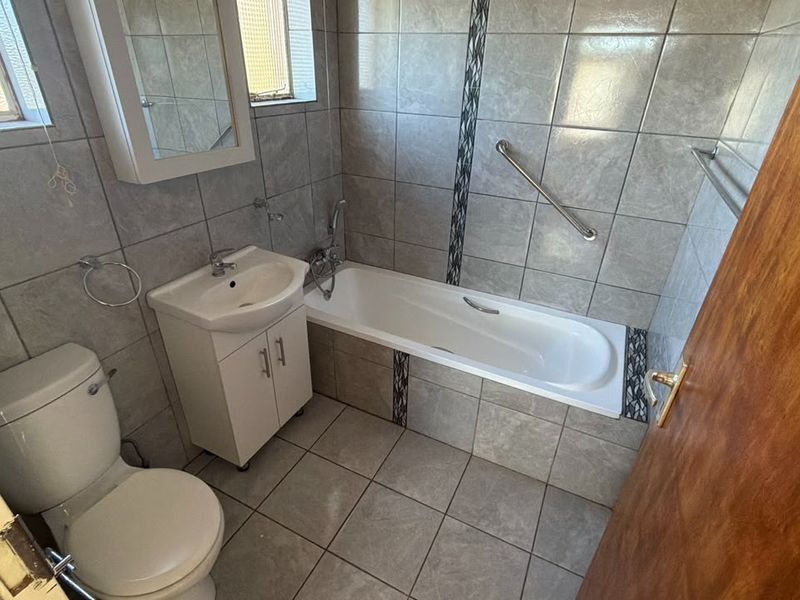 A functional bathroom featuring tiled walls and flooring, a built-in vanity with a washbasin, a toilet, and a bathtub with a safety grab rail.