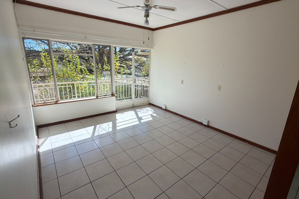 A bright, empty living room featuring tiled flooring, a ceiling fan, and large windows with integrated security bars, offering natural light and garden views.
