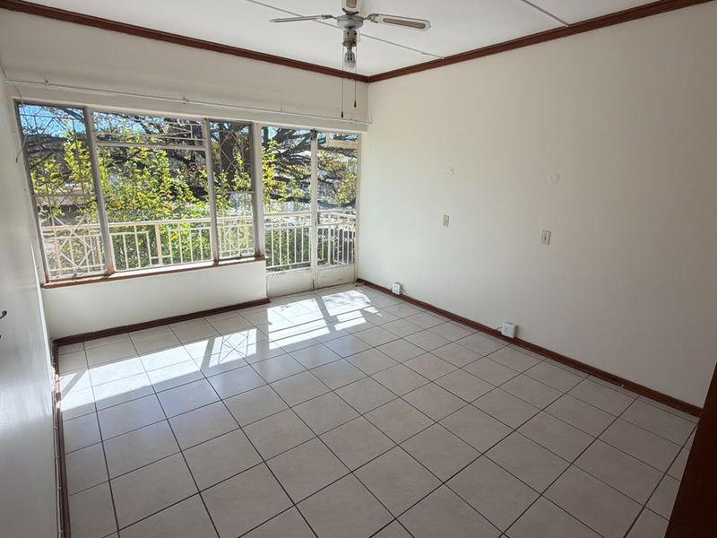 A bright, empty living room featuring tiled flooring, a ceiling fan, and large windows with integrated security bars, offering natural light and garden views.