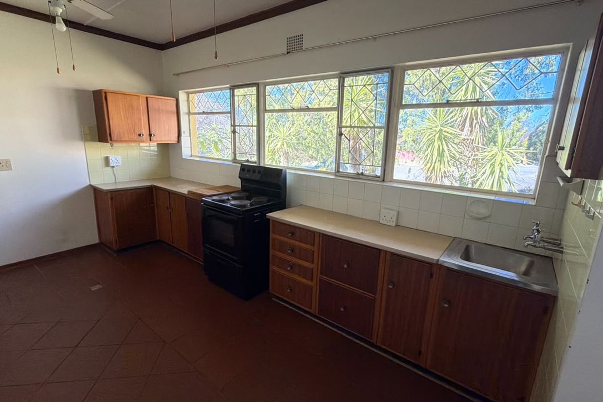 A dated, empty kitchen featuring original wooden cabinetry, laminate countertops, a freestanding stove, and large leaded glass windows overlooking the garden.