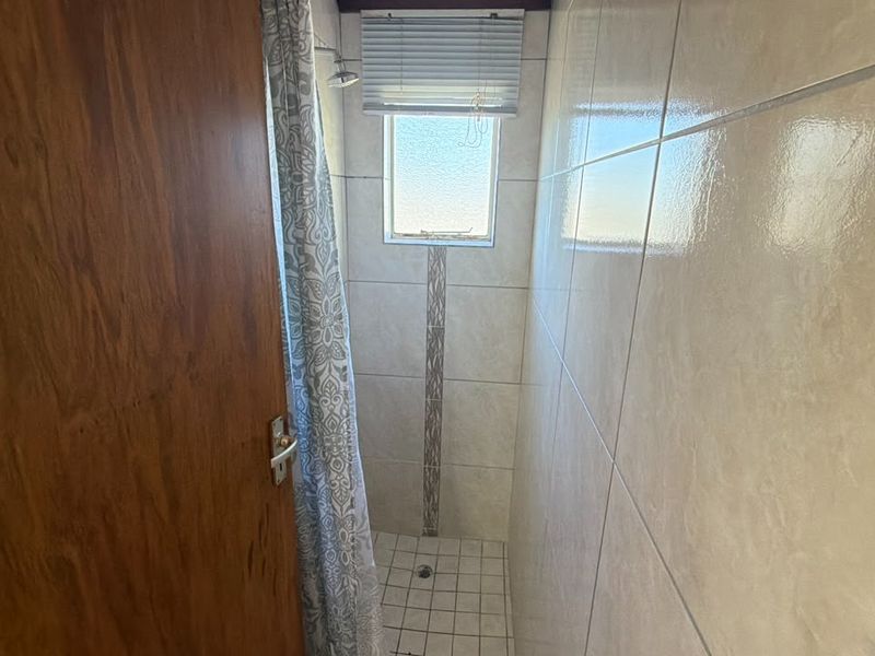A functional shower enclosure featuring neutral wall tiles, a tiled floor, and a small window with a blind.