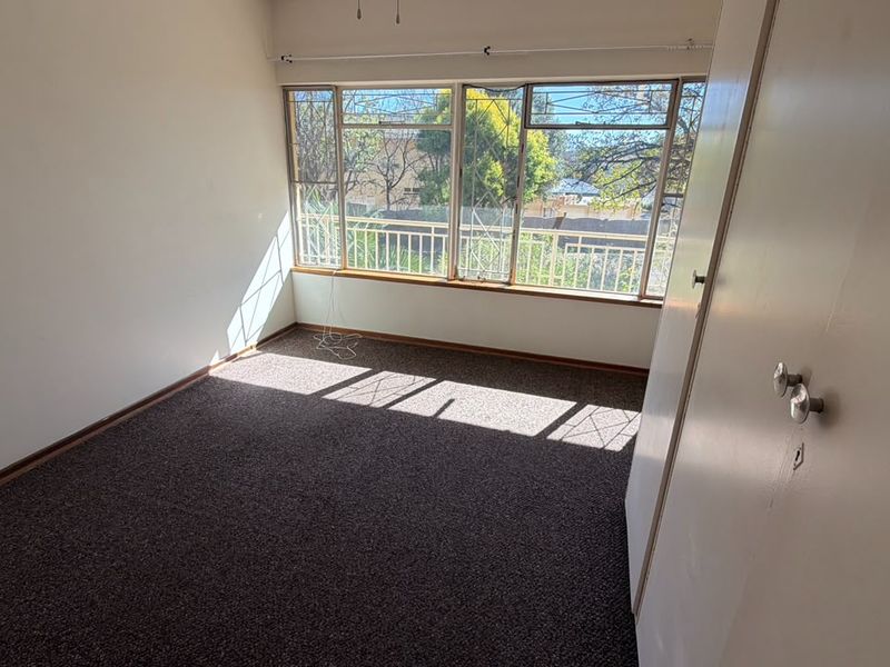 A bright, empty bedroom featuring carpeted flooring, a built-in wardrobe, and large windows with security bars.