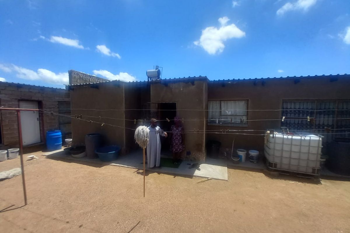 Rear exterior view of a single-story dwelling featuring a solar water heater and basic masonry construction with a dirt yard.