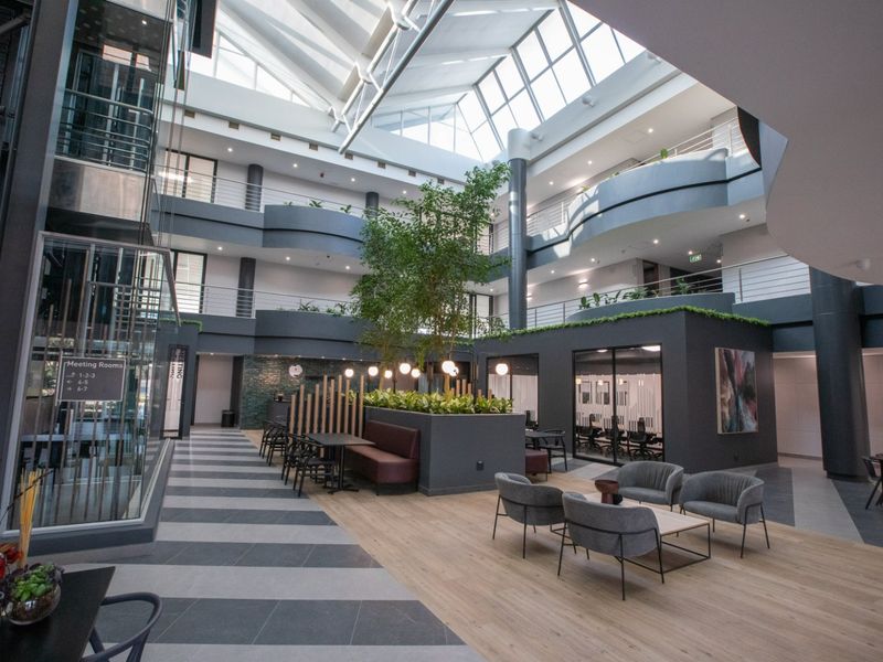 Modern commercial atrium featuring a soaring skylight, multi-level mezzanine walkways, glass elevator, and a central indoor garden area with integrated seating.