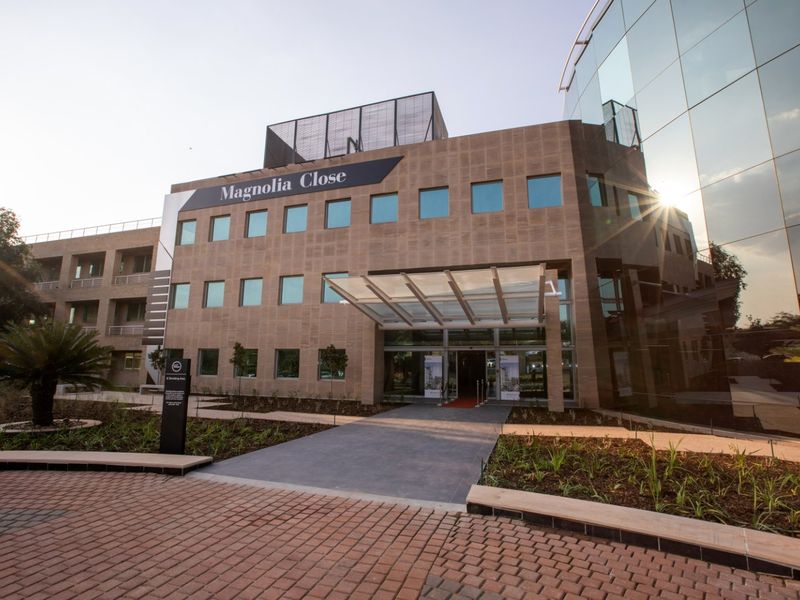 Modern commercial office building facade featuring stone cladding, extensive glass curtain walling, and a prominent glass entrance canopy with landscaped surroundings.