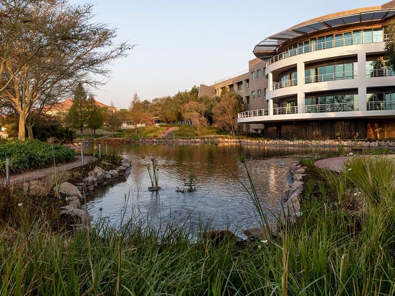 A scenic view of a modern commercial building featuring multi-level balconies overlooking a landscaped garden with a central pond and paved walkways.