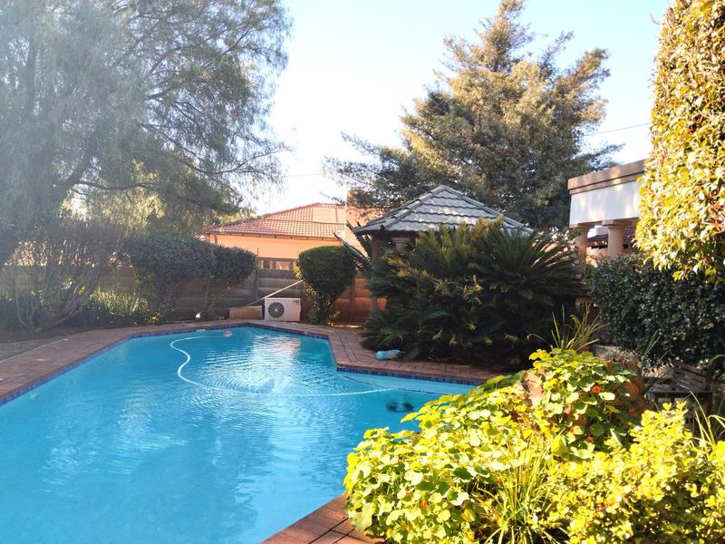 A well-maintained outdoor swimming pool area featuring brick coping, lush garden landscaping, and a covered gazebo structure.