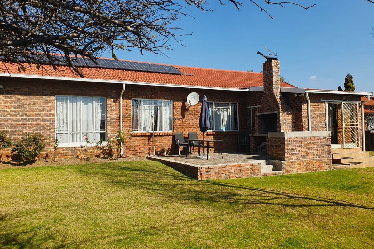 Rear view of a brick house featuring a paved patio with a built-in brick braai, solar panels on the roof, and a well-maintained garden.