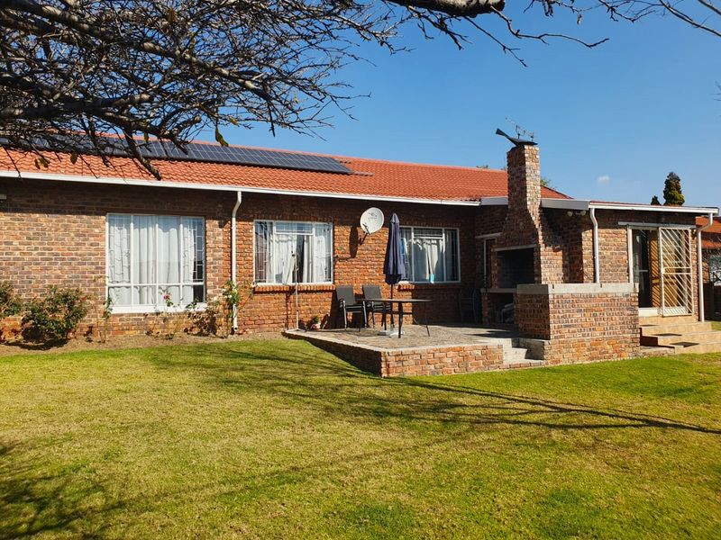 Rear view of a brick house featuring a paved patio with a built-in brick braai, solar panels on the roof, and a well-maintained garden.