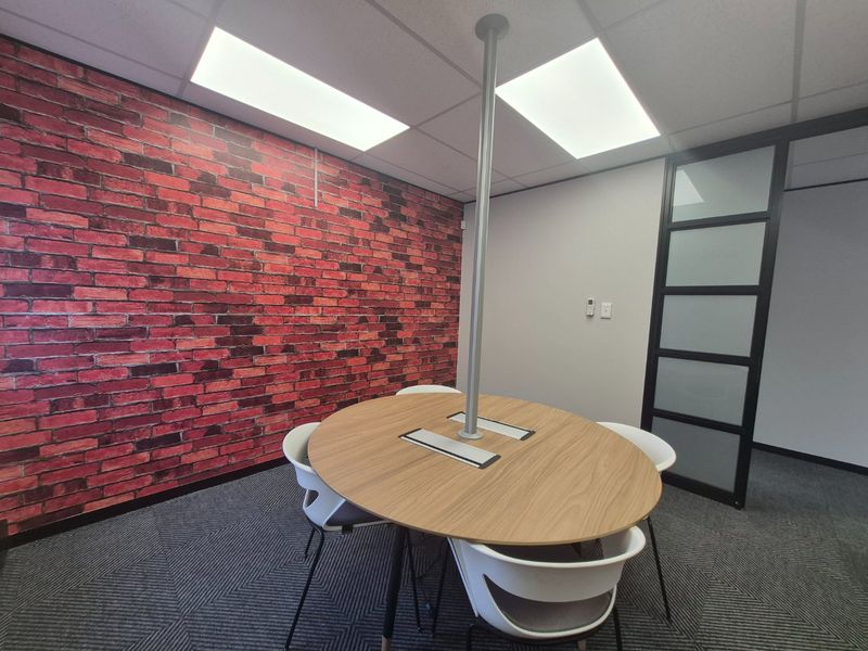 A modern office meeting room featuring a central table, glass partition wall, carpet tiles, and a decorative brick-effect feature wall.