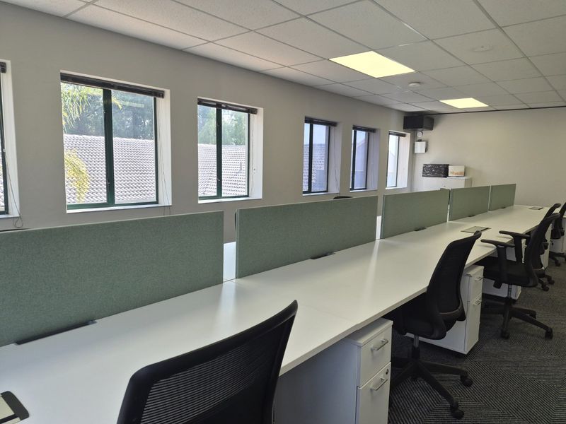 A professional commercial office space featuring a row of built-in workstations with privacy partitions, carpeted flooring, and ample natural light from multiple windows.