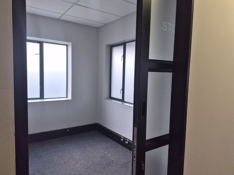 A clean, empty office space featuring carpet tiles, a suspended ceiling, and a modern sliding glass door with natural light from two windows.