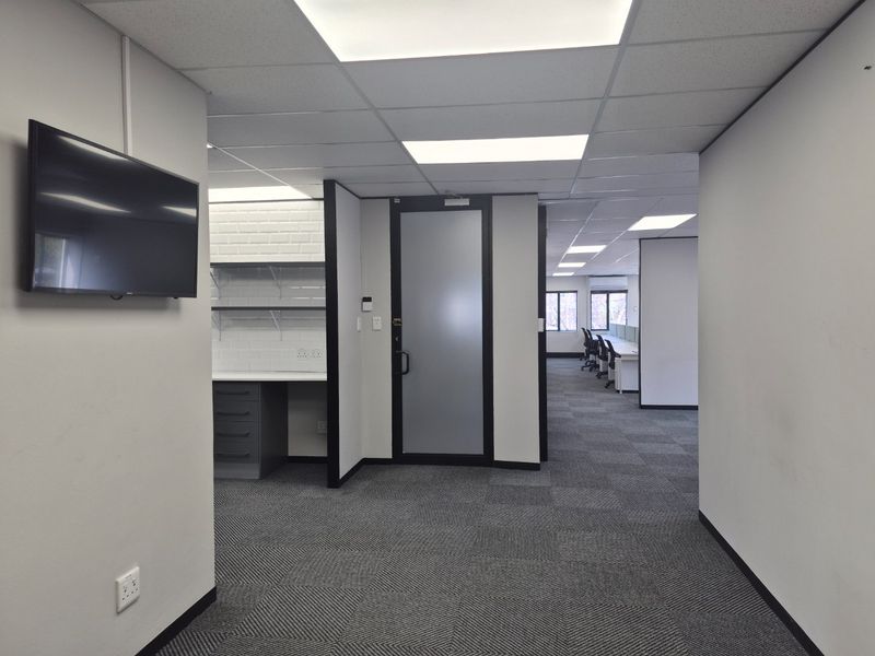 A modern, professional office space featuring carpeted flooring, a suspended ceiling with recessed lighting, and a dedicated kitchenette area with built-in cabinetry.
