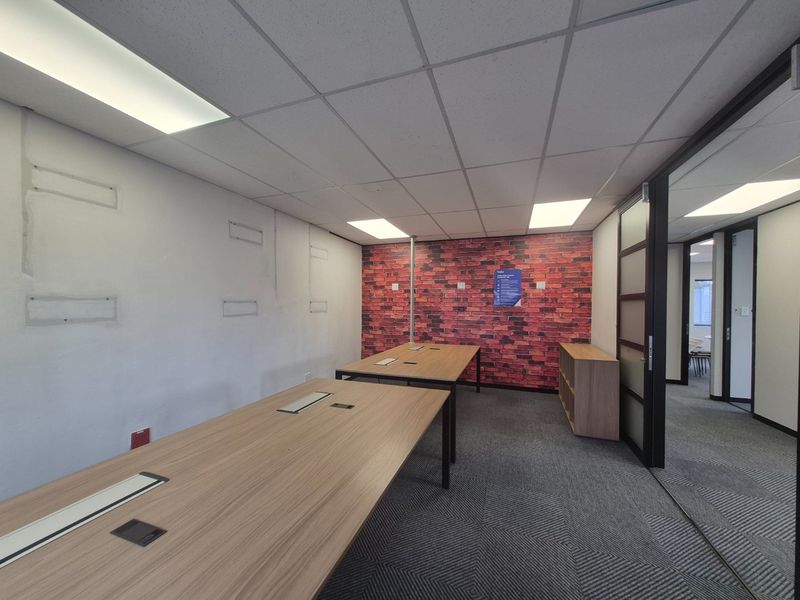 A functional office space featuring a brick-effect accent wall, suspended ceiling with recessed lighting, carpeted flooring, and integrated desk workstations.