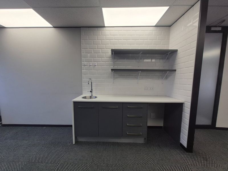 A clean, modern utility kitchenette area featuring dark grey cabinetry, a stainless steel sink, subway tile backsplash, and open shelving in a professional office setting.