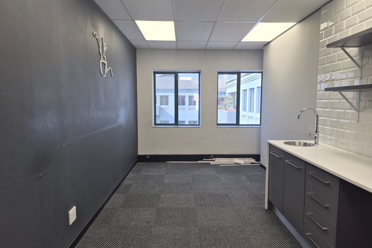 A modern, empty office kitchenette featuring dark cabinetry, a subway tile backsplash, stainless steel sink, and carpeted flooring.