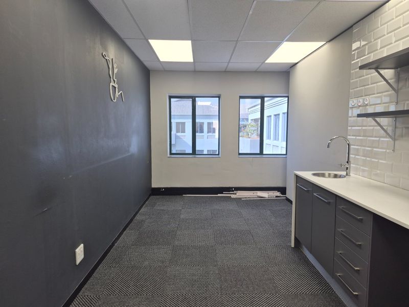 A modern, empty office kitchenette featuring dark cabinetry, a subway tile backsplash, stainless steel sink, and carpeted flooring.