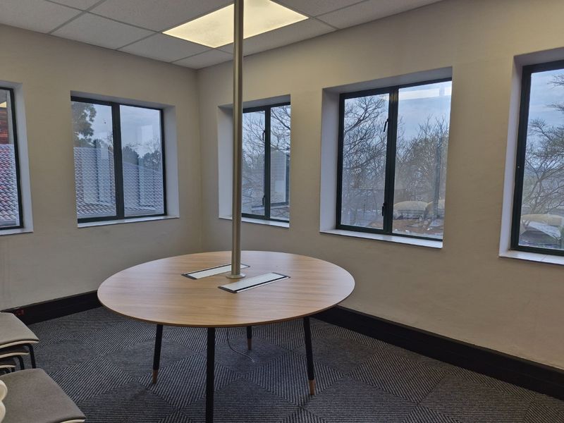 A bright, modern office space featuring large windows, patterned carpet tiles, and a central meeting table with integrated power connectivity.
