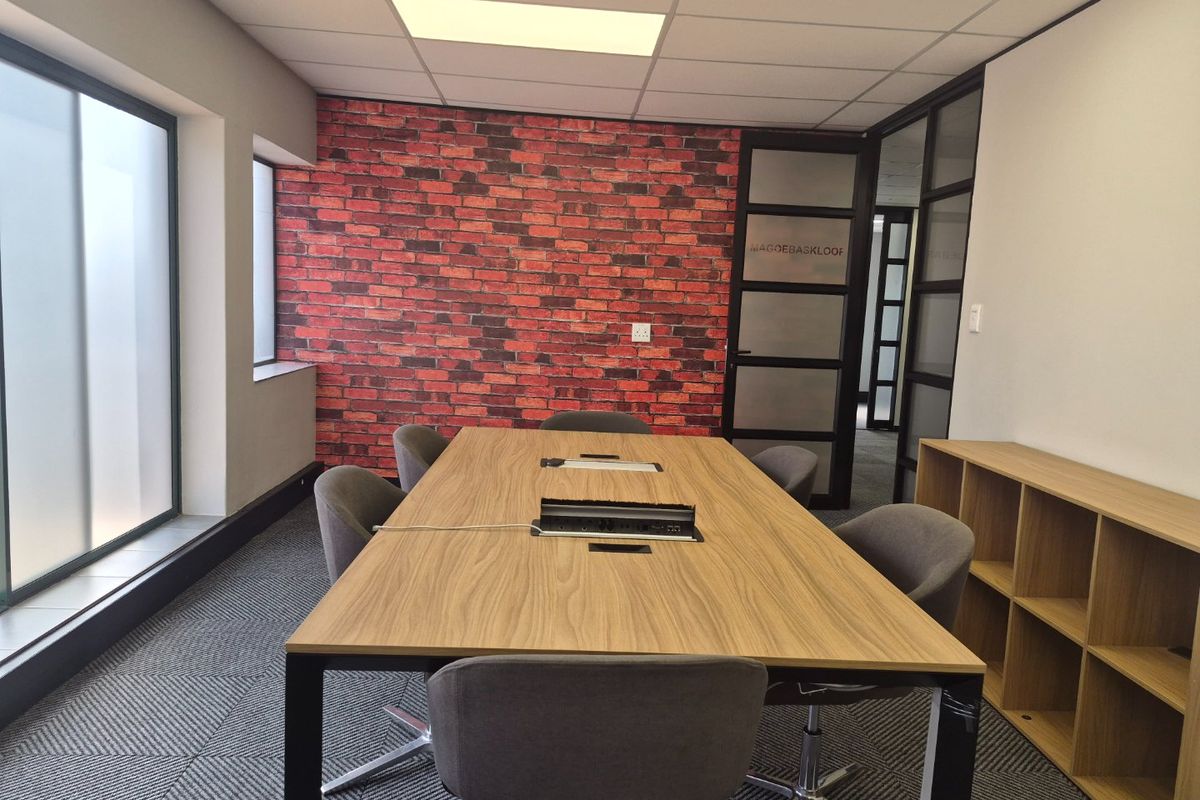 A modern office meeting room featuring a brick accent wall, suspended ceiling, glass partition doors, and built-in shelving.