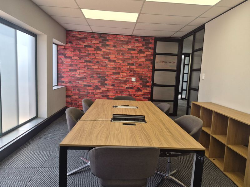 A modern office meeting room featuring a brick accent wall, suspended ceiling, glass partition doors, and built-in shelving.