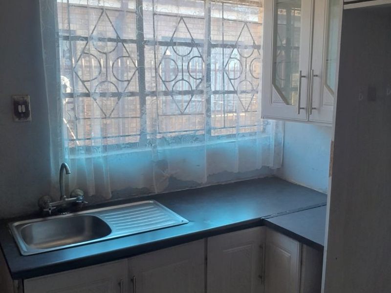 A compact kitchen area featuring built-in cabinetry, a stainless steel sink, and security bars on the window, requiring cosmetic updates to improve its condition.