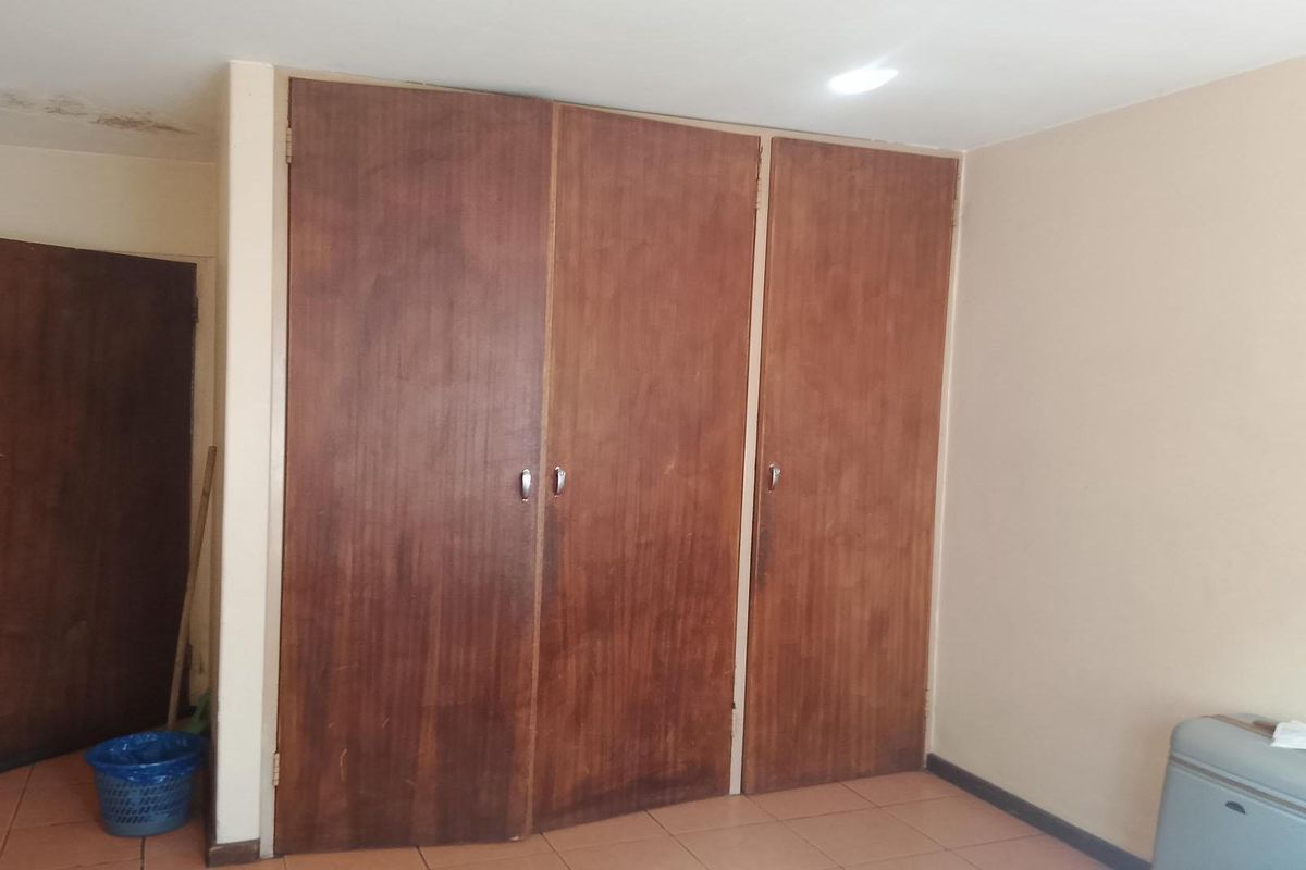 A bedroom featuring built-in wooden wardrobes, tiled flooring, and recessed lighting, showing signs of ceiling water damage and dated finishes.