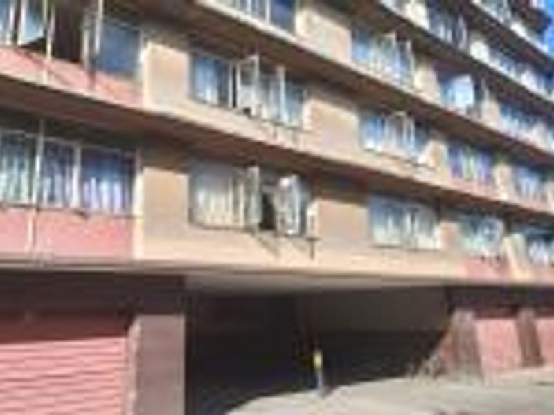 Exterior view of a multi-story apartment building featuring a ground-level parking area with roller doors and multiple casement windows along the facade.