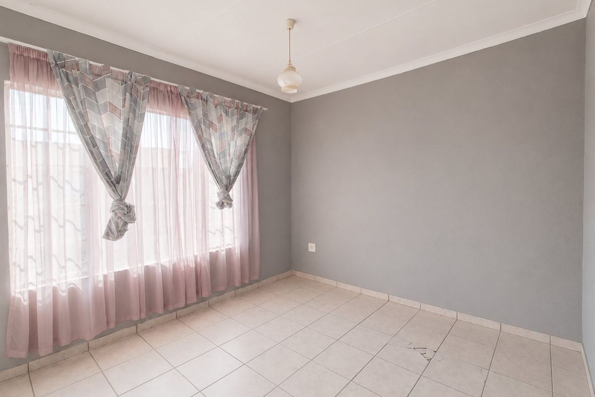 A bright, empty bedroom featuring neutral grey walls, tiled flooring, and a large window with curtains.