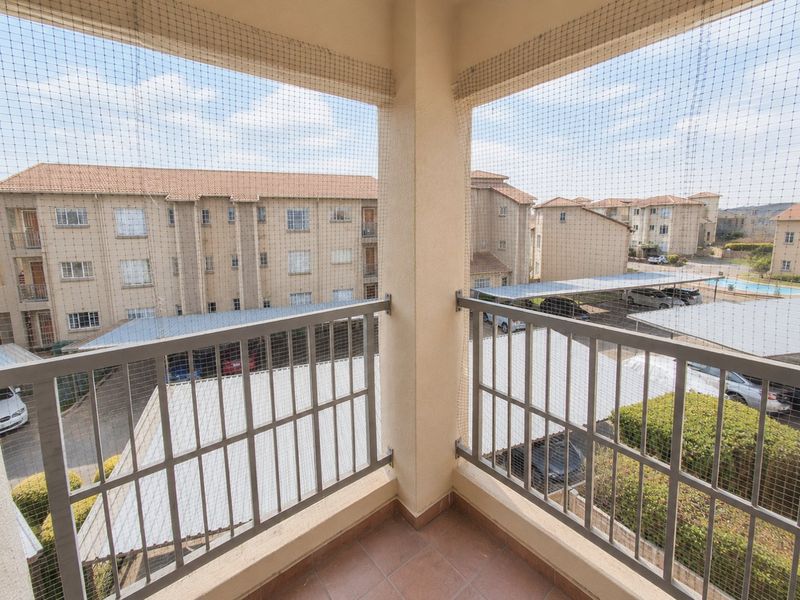 A covered balcony featuring tiled flooring and a metal balustrade with safety netting, overlooking the apartment complex parking area.
