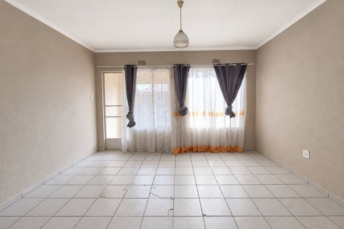 An empty living room featuring neutral walls, tiled flooring with visible cracks, and natural light through large windows and a sliding door.