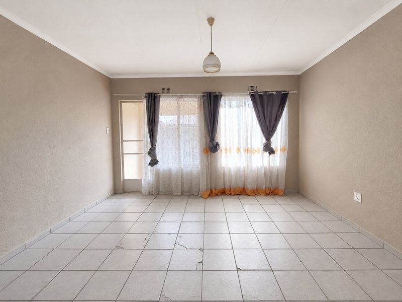 An empty living room featuring neutral walls, tiled flooring with visible cracks, and natural light through large windows and a sliding door.