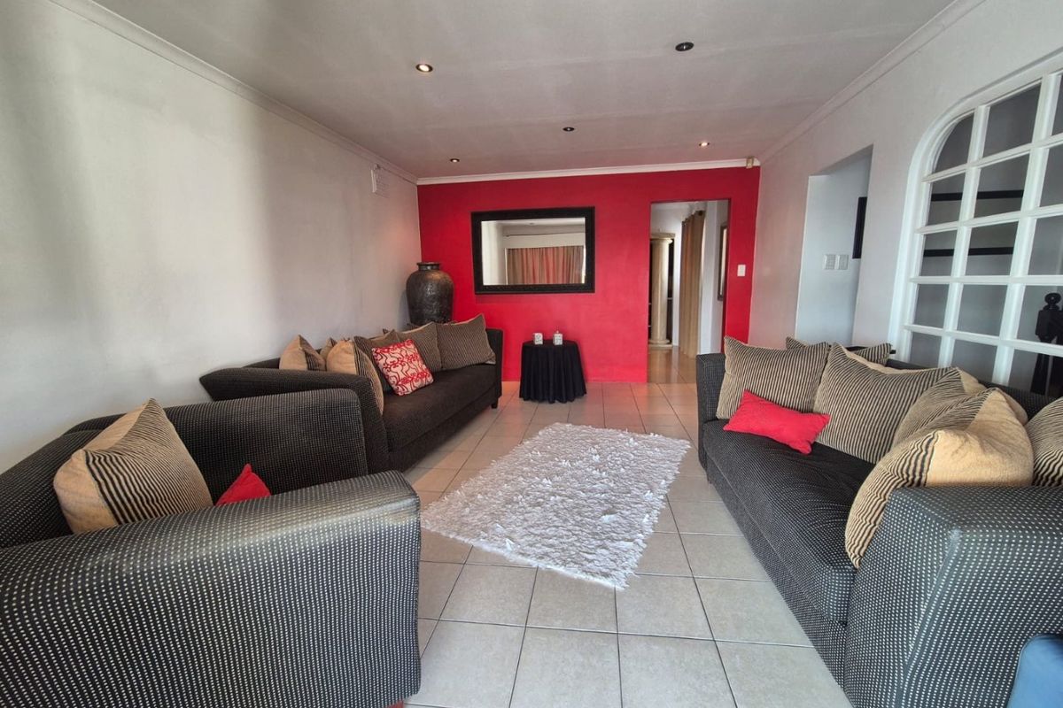 A spacious living room featuring neutral ceramic floor tiles, recessed downlighting, and a bold red accent wall with an arched doorway leading to other areas.