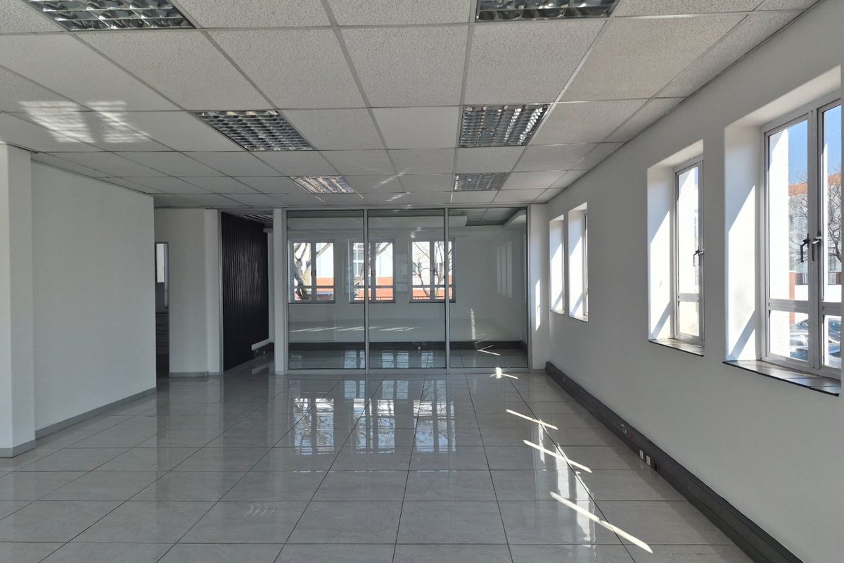 A bright, vacant commercial office space featuring polished tiled floors, a suspended ceiling with recessed lighting, and large windows providing ample natural light.