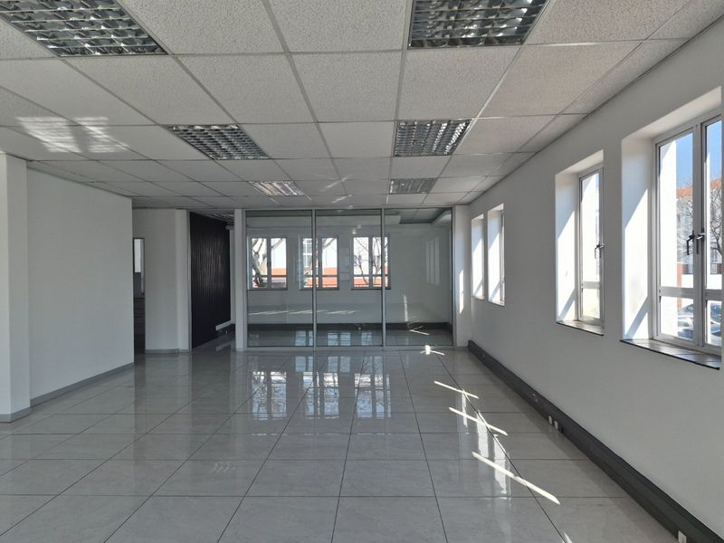 A bright, vacant commercial office space featuring polished tiled floors, a suspended ceiling with recessed lighting, and large windows providing ample natural light.