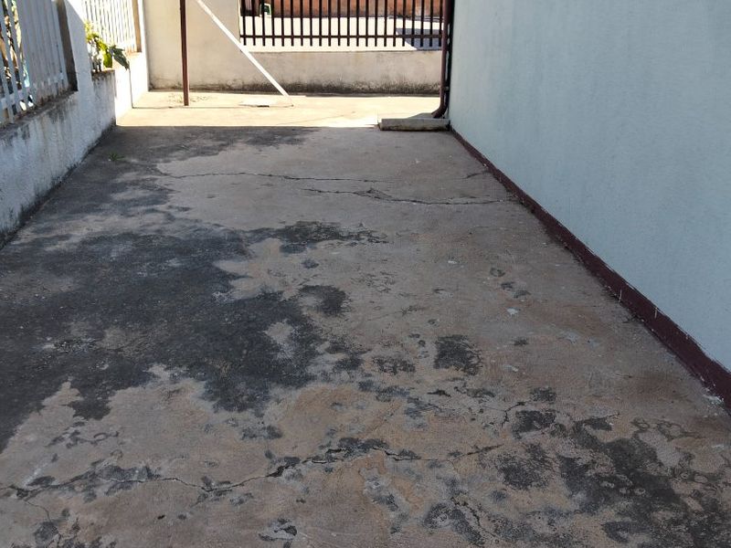 A side or rear exterior area featuring concrete paving, metal perimeter fencing, and a downpipe, showing signs of wear and surface cracking.