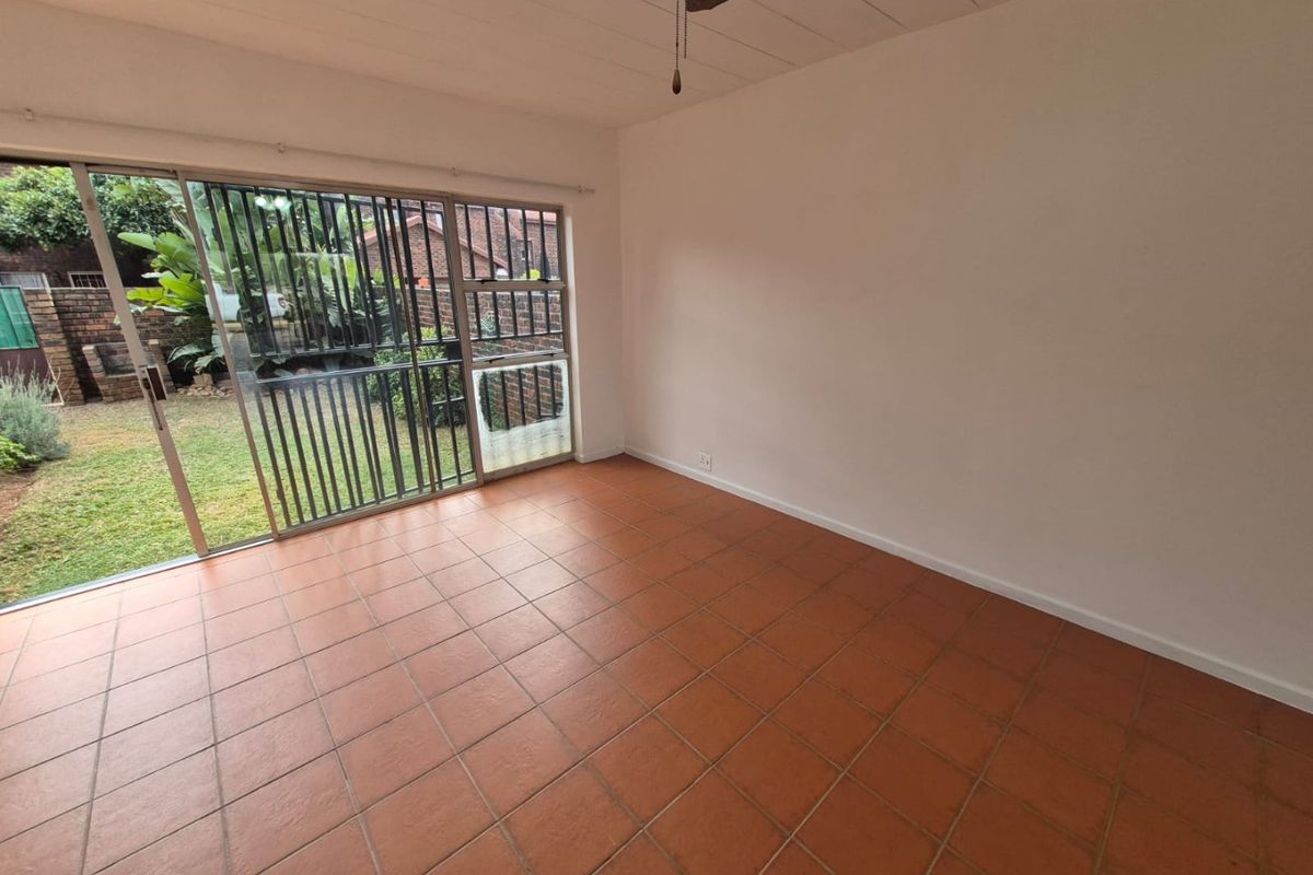 An empty living room featuring terracotta floor tiles, a ceiling fan, and a large sliding glass door with a security gate leading to the garden.