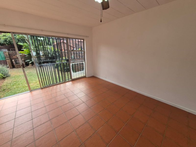 An empty living room featuring terracotta floor tiles, a ceiling fan, and a large sliding glass door with a security gate leading to the garden.