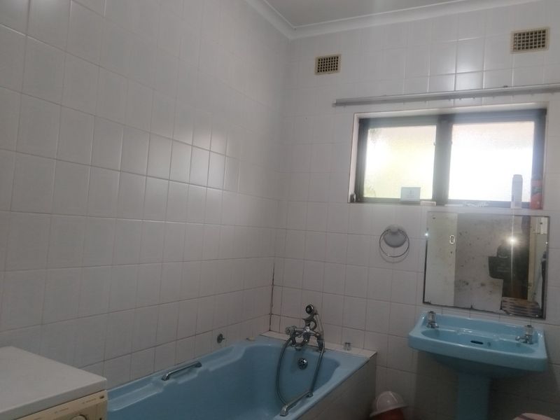 A dated bathroom featuring a blue bathtub and matching pedestal basin, with white wall tiles and a window for natural light.