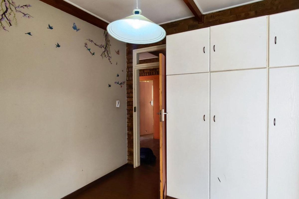 A bedroom featuring built-in cupboards, exposed timber ceiling beams, and an exposed brick wall, presenting a dated interior in need of cosmetic updates.